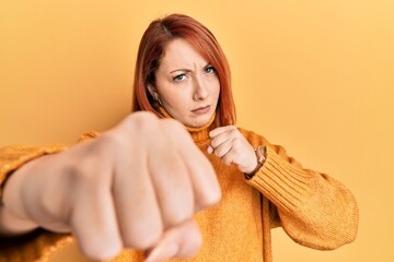 Beautiful redhead woman wearing casual winter sweater over yellow background punching fist to...