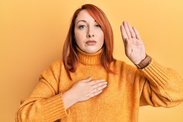Beautiful redhead woman wearing casual winter sweater over yellow background swearing with hand on...