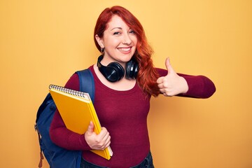 Young beautiful redhead student woman wearing backpack and headphones holding notebook smiling happy and positive, thumb up doing excellent and approval sign