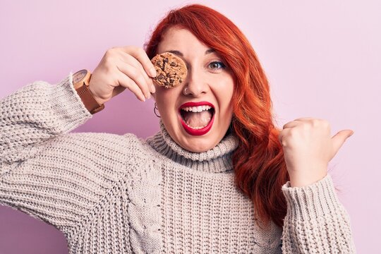 Young beautiful redhead woman standing holding chocolate cookie over eye pointing thumb up to the side smiling happy with open mouth