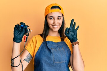 Young hispanic woman tattoo artist wearing professional uniform and gloves holding tattooer machine...