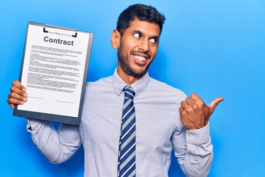 Young latin man holding clipboard with contract document pointing thumb up to the side smiling happy with open mouth