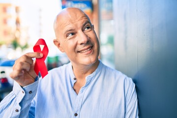 Middle age bald man smiling happy holding red hiv ribbon leaning on the wall at the city