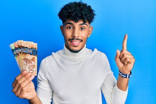 Young arab handsome man holding canadian dollars smiling with an idea or question pointing finger with happy face, number one