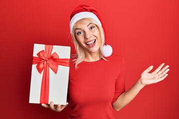 Young blonde girl wearing christmas hat and holding a gift celebrating achievement with happy smile and winner expression with raised hand
