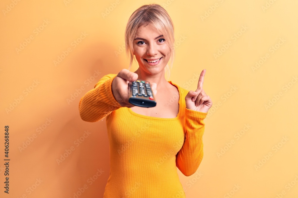 Poster blonde woman changing tv channel using television remote control over yellow background smiling with