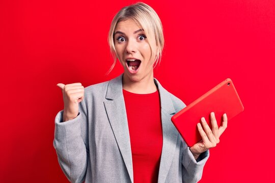 Young beautiful blonde businesswoman working using tablet over isolated red background pointing thumb up to the side smiling happy with open mouth
