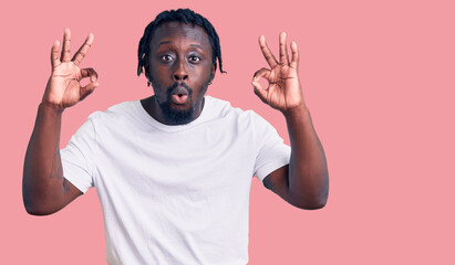 Fototapeta premium Young african american man with braids wearing casual white tshirt looking surprised and shocked doing ok approval symbol with fingers. crazy expression
