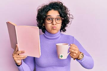 Young hispanic woman with curly hair reading a book and drinking a cup of coffee puffing cheeks with funny face. mouth inflated with air, catching air.