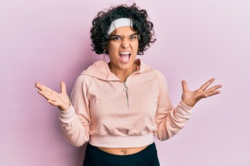 Young hispanic woman with curly hair wearing sportswear crazy and mad shouting and yelling with aggressive expression and arms raised. frustration concept.