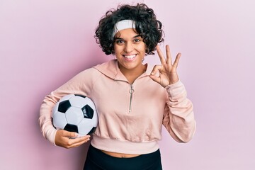 Young hispanic woman with curly hair holding soccer ball doing ok sign with fingers, smiling friendly gesturing excellent symbol