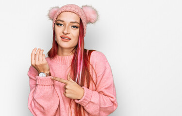 Young caucasian woman wearing casual clothes and wool cap in hurry pointing to watch time, impatience, looking at the camera with relaxed expression