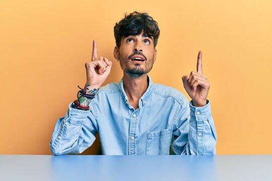 Young hispanic man wearing casual clothes sitting on the table amazed and surprised looking up and pointing with fingers and raised arms.
