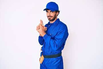 Handsome young man with curly hair and bear weaing handyman uniform holding symbolic gun with hand gesture, playing killing shooting weapons, angry face