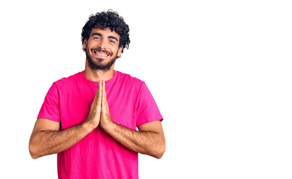 Handsome young man with curly hair and bear wearing casual pink tshirt praying with hands together asking for forgiveness smiling confident.