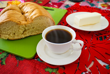 A white cup with hot black coffee, on a table with a red Christmas tablecloth, behind a Dominican telera bread and a stick of butter.