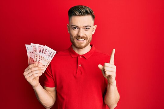 Young redhead man holding 100 new zealand dollars banknote smiling with an idea or question pointing finger with happy face, number one