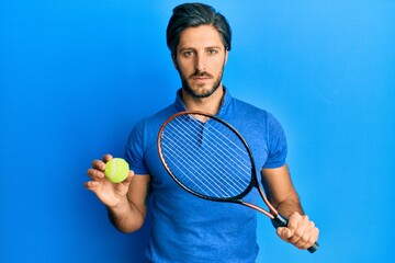 Young hispanic man playing tennis holding racket and ball relaxed with serious expression on face....