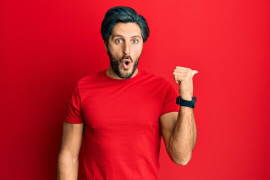 Young hispanic man wearing casual red t shirt surprised pointing with hand finger to the side, open mouth amazed expression.
