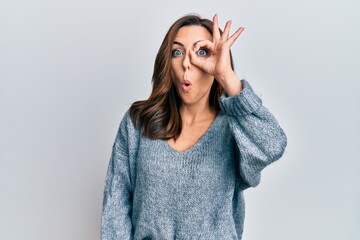 Young brunette woman wearing casual winter sweater doing ok gesture shocked with surprised face, eye looking through fingers. unbelieving expression.