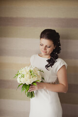 Pretty young Bride in simple wedding dress with her wedding bouquet. Black-haired woman with wedding hair-style with a long tail. Boudoir morning of the bride in modern room