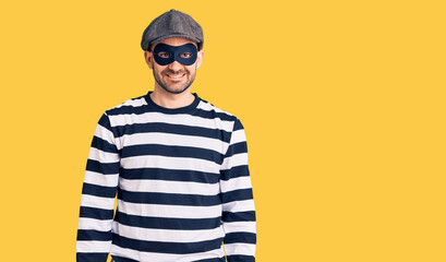 Young handsome man wearing burglar mask with a happy and cool smile on face. lucky person.