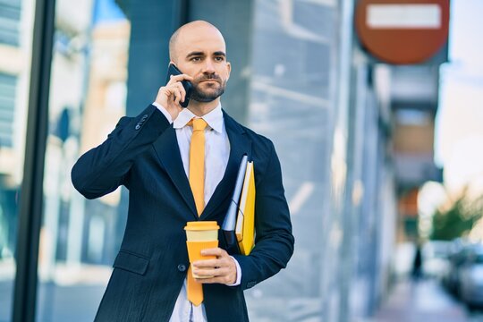 Young hispanic bald businessman with serious expression talking on the smartphone drinking coffee at the city