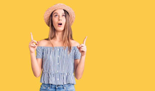 Young beautiful girl wearing hat and t shirt amazed and surprised looking up and pointing with fingers and raised arms.