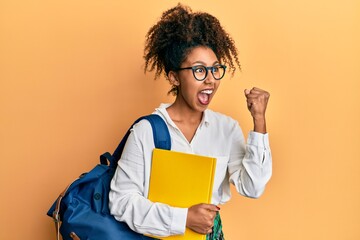 Beautiful african american woman with afro hair wearing school bag and holding books screaming proud, celebrating victory and success very excited with raised arms