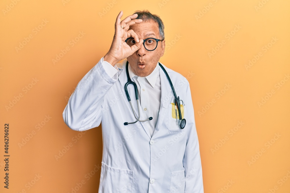 Canvas Prints Middle age indian man wearing doctor coat and stethoscope doing ok gesture shocked with surprised face, eye looking through fingers. unbelieving expression.