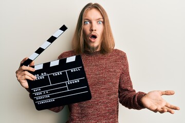 Handsome caucasian man with long hair holding video film clapboard scared and amazed with open...