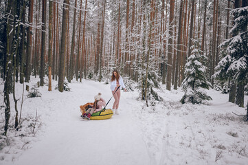 Happy mother and little cute girl in pink warm outwear walking having fun rides inflatable snow tube with red shiba inu dog in snowy white winter forest outdoors. Family sport vacation activities