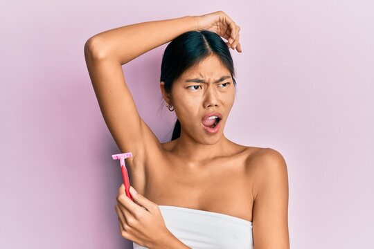 Young chinese woman wearing shower towel using razor angry and mad screaming frustrated and furious, shouting with anger. rage and aggressive concept.