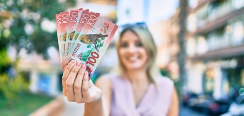 Young blonde woman smiling happy holding new zealand 100 dollars banknotes at the city.