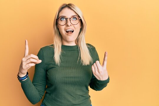 Young caucasian woman wearing casual clothes and glasses amazed and surprised looking up and pointing with fingers and raised arms.