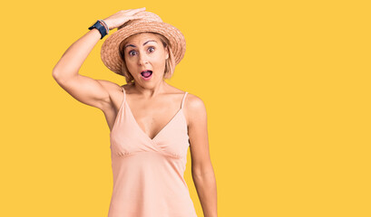 Young blonde woman wearing summer hat surprised with hand on head for mistake, remember error....