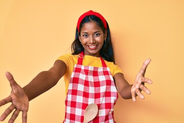 Young indian girl wearing baker apron looking at the camera smiling with open arms for hug. cheerful expression embracing happiness.