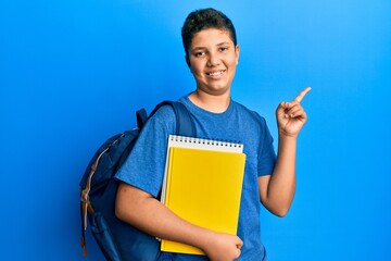 Teenager hispanic boy wearing school bag and holding books smiling happy pointing with hand and...