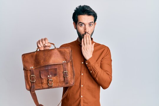 Young hispanic man wearing leather bag covering mouth with hand, shocked and afraid for mistake. surprised expression