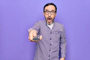 Middle age man changing tv channel using television remote control over purple background scared...