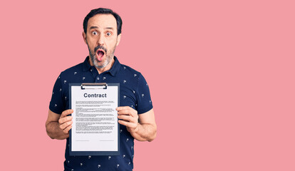Middle age handsome man holding clipboard with contract document scared and amazed with open mouth...