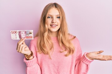 Beautiful young caucasian girl holding 500 swedish krona banknote celebrating achievement with happy smile and winner expression with raised hand