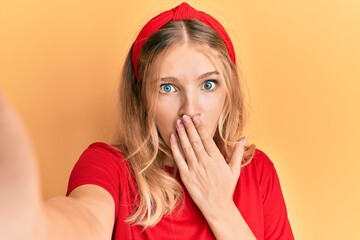 Beautiful young caucasian girl taking a selfie photo covering mouth with hand, shocked and afraid...