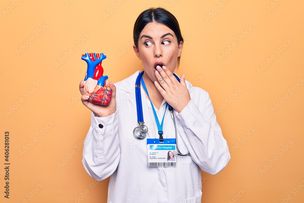 Canvas Prints Young beautiful cardiologist woman wearing stethoscope holding heart over yellow background covering mouth with hand, shocked and afraid for mistake. Surprised expression