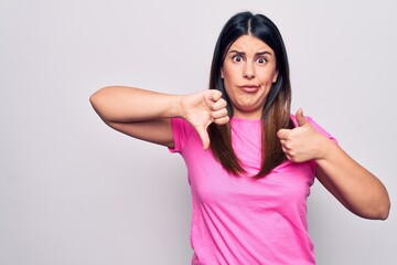 Obraz na płótnie Canvas Young beautiful brunette woman wearing casual pink t-shirt standing over white background Doing thumbs up and down, disagreement and agreement expression. Crazy conflict