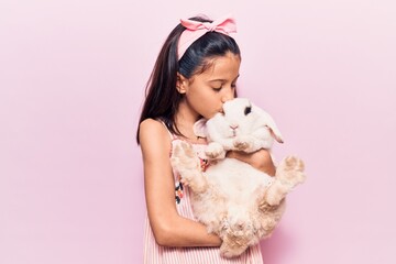 Adorable hispanic kid girl wearing diadem smiling happy. Holding and kissing cute rabbit standing over isolated pink background