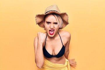 Young beautiful blonde woman wearing bikini and summer hat angry and mad raising fist frustrated and furious while shouting with anger. rage and aggressive concept.