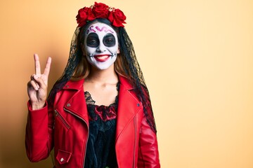Woman wearing day of the dead costume over yellow smiling with happy face winking at the camera doing victory sign. number two.
