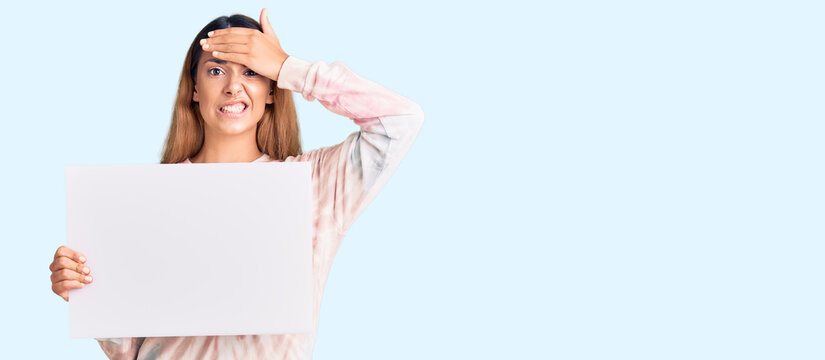Beautiful young woman holding blank empty banner stressed and frustrated with hand on head, surprised and angry face