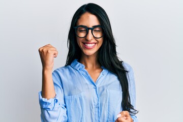 Beautiful hispanic woman wearing business clothes and glasses celebrating surprised and amazed for success with arms raised and eyes closed